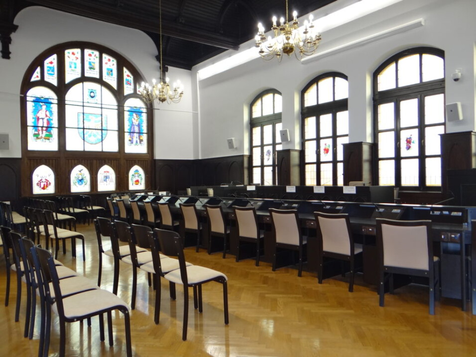 Sala obrad z&nbsp;rzędami krzeseł, długim stołem prezydialnym i&nbsp;ozdobnymi witrażami w&nbsp;oknach.